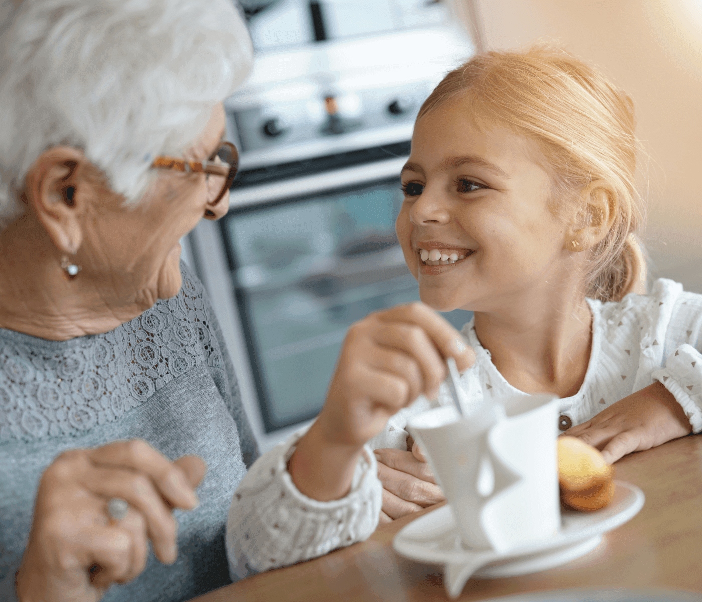Elderly woman and young girl smiling at each other while having tea and pastries at a table in a home kitchen. - Home Instead
