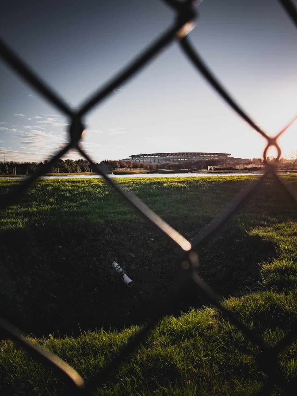 Chain-link fence framing a distant stadium under a clear sky, with grass and sunlight in the foreground. - Home Instead