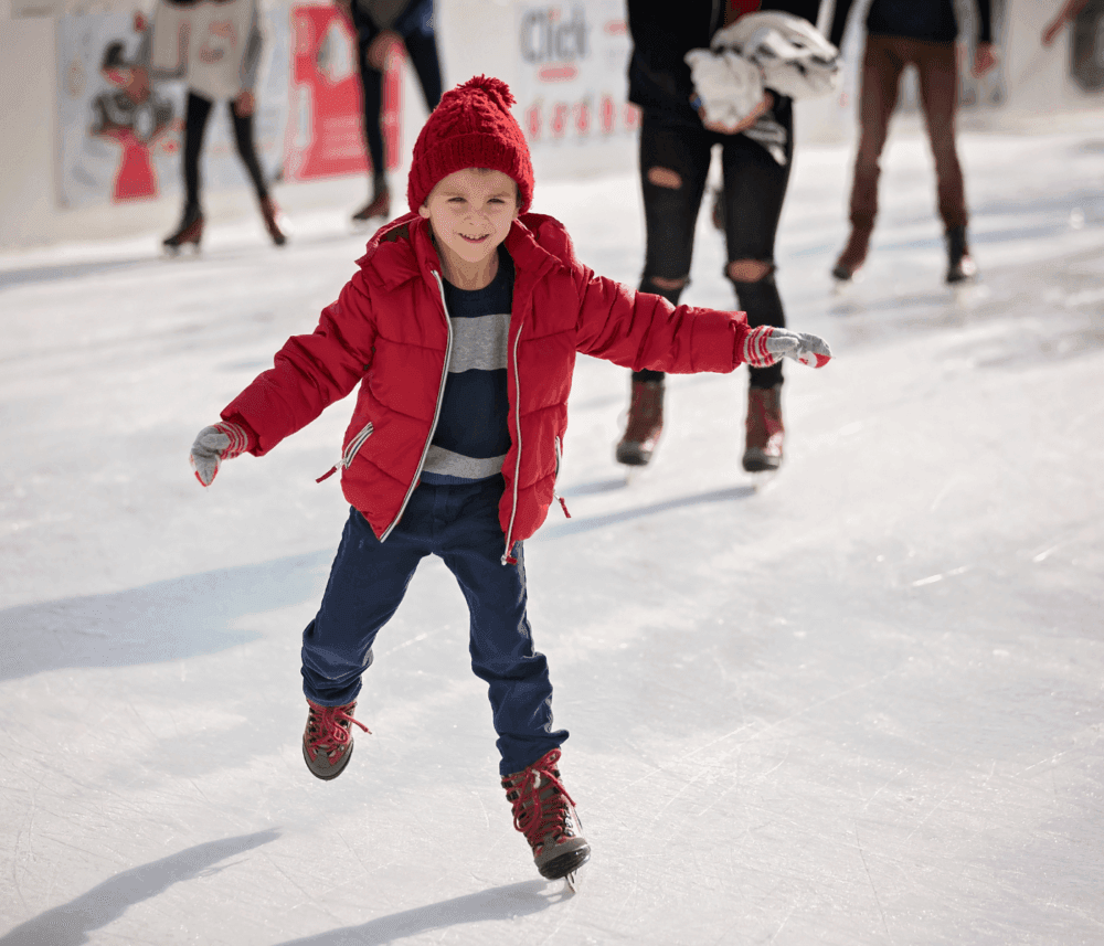 A child in a red jacket and hat happily ice skating on a rink, with other skaters in the background. - Home Instead