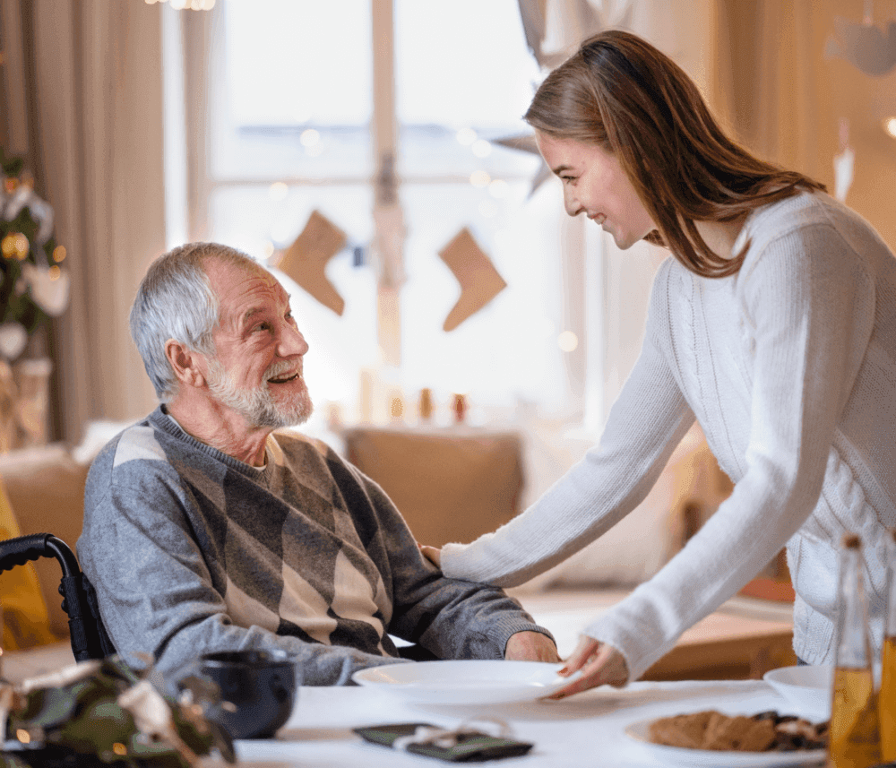 Young woman serving a smiling elderly man in a wheelchair at a holiday-decorated table. - Home Instead
