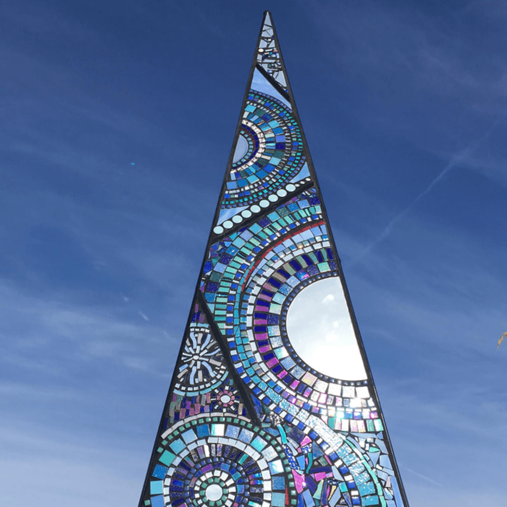 A tall, triangular stained glass sculpture against a clear blue sky, with intricate geometric patterns in various colors. - Home Instead