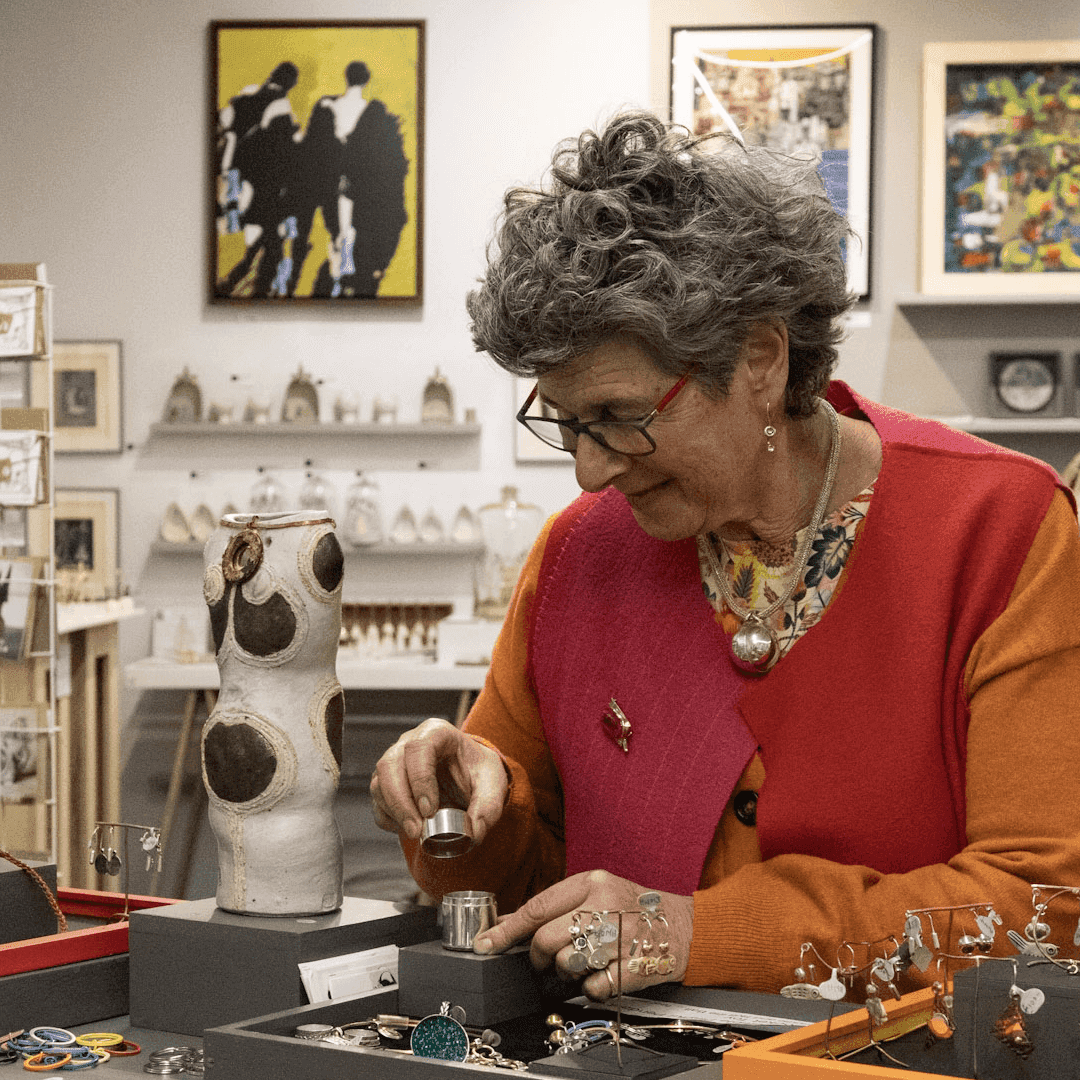 A woman with curly hair examines jewelry in a brightly decorated shop filled with various art pieces. - Home Instead