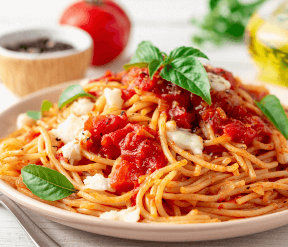 A plate of spaghetti topped with tomato sauce, cheese, and fresh basil leaves, with a tomato and spices in the background. - Home Instead