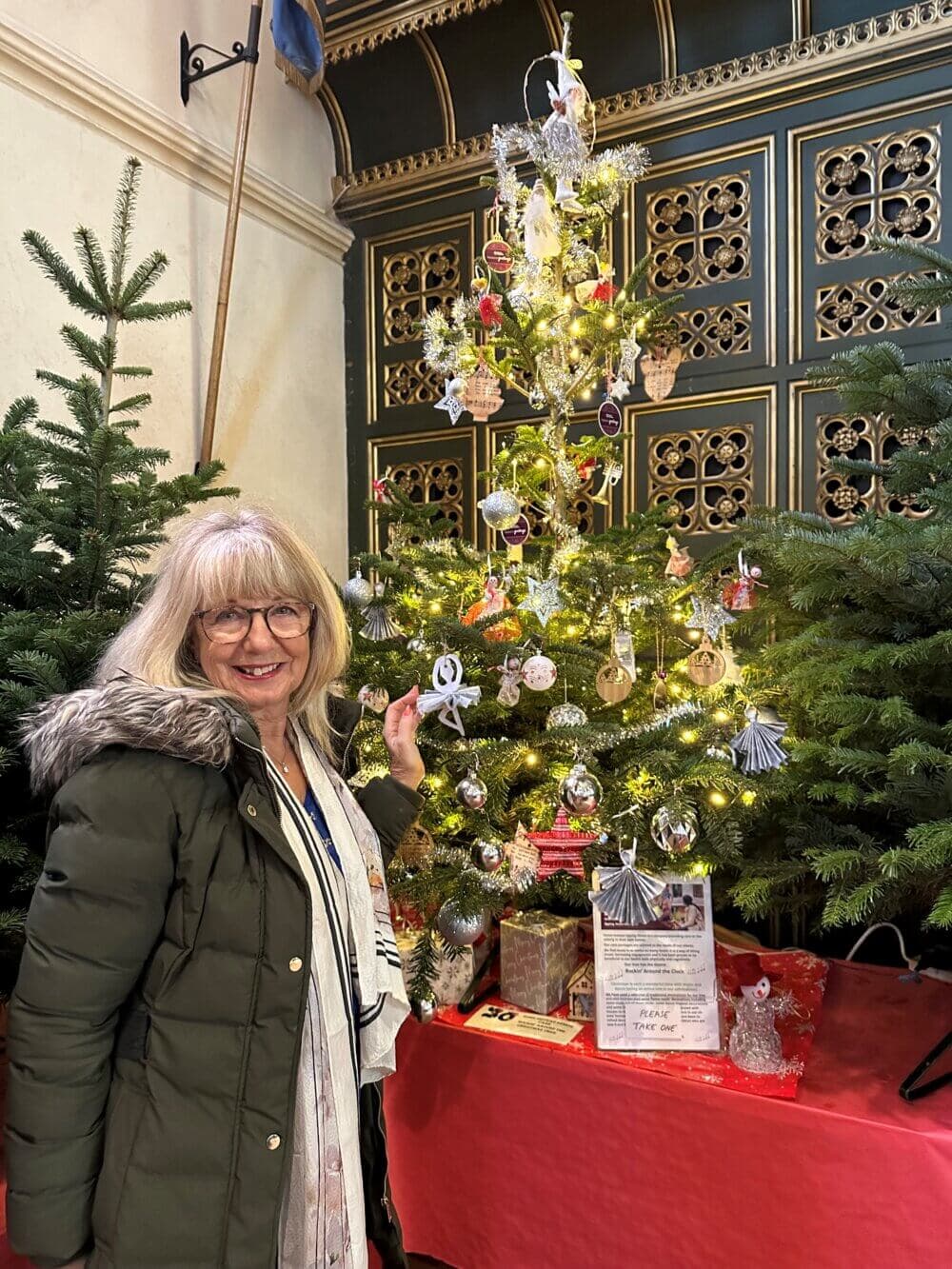 A woman in a green coat smiles while standing next to a decorated Christmas tree indoors. - Home Instead