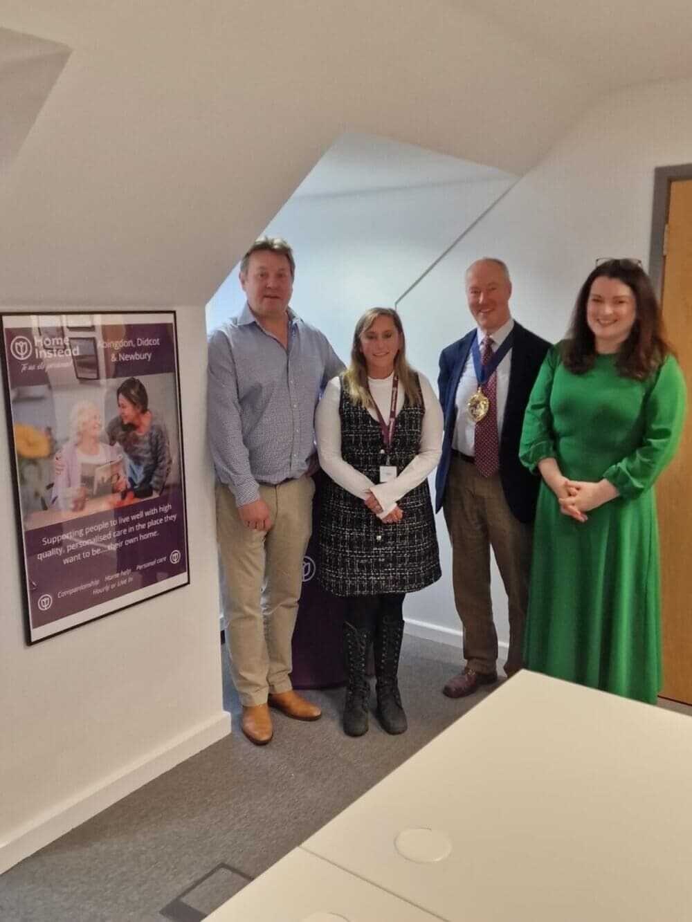 Four people standing in a room near a sign with text, smiling at the camera. One is wearing a ceremonial chain. - Home Instead