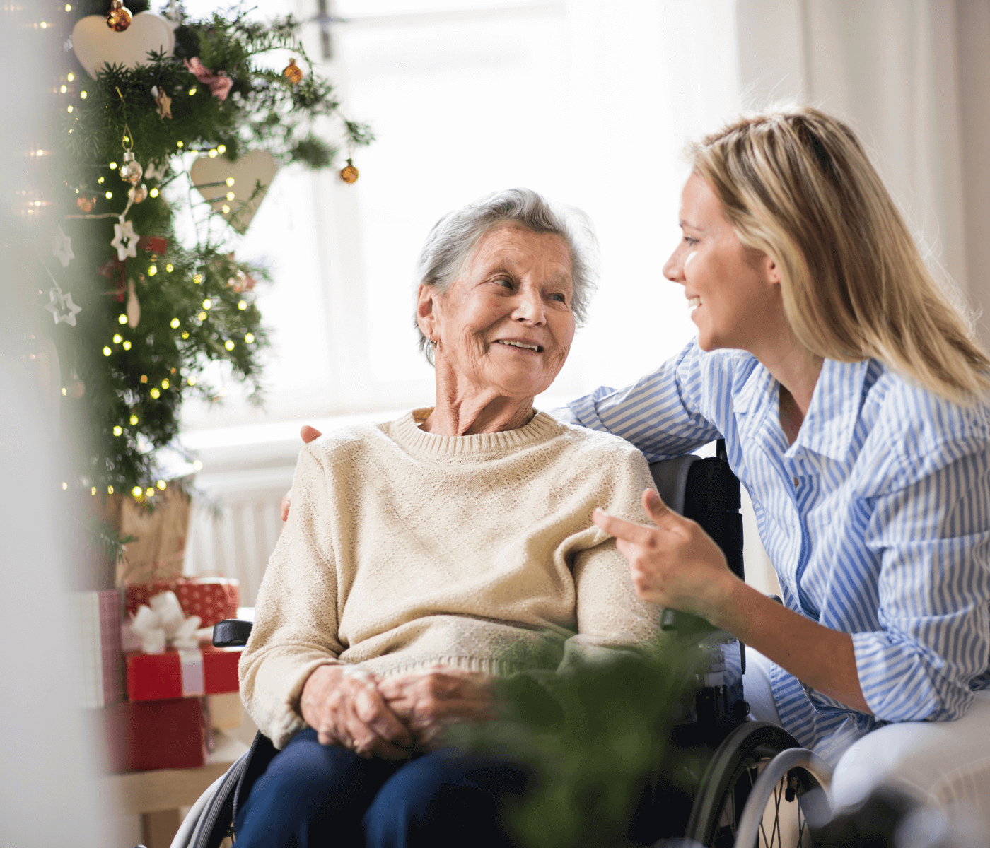 A younger woman smiles and talks to an elderly woman in a wheelchair near a decorated Christmas tree. - Home Instead