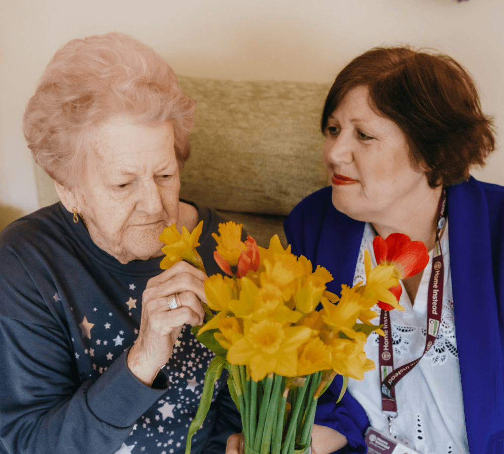 An elderly woman holding a bouquet of yellow flowers while a caregiver sits beside her, both smiling warmly. - Home Instead
