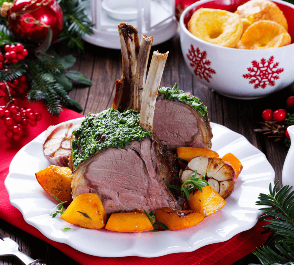 Roast rack of lamb with herbs, served with roasted vegetables on a white plate, holiday decor in the background. - Home Instead