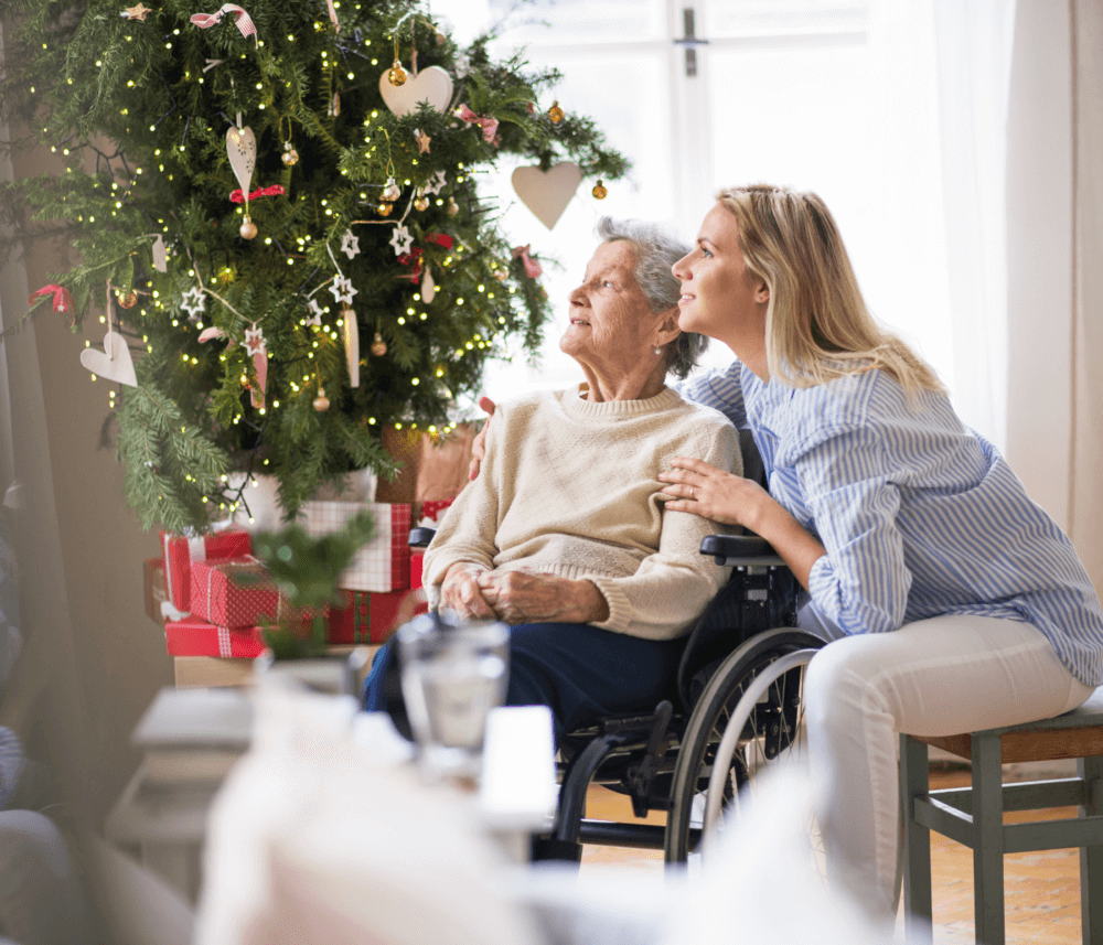 A young woman and an elderly woman in a wheelchair look at a decorated Christmas tree with gifts below it. - Home Instead