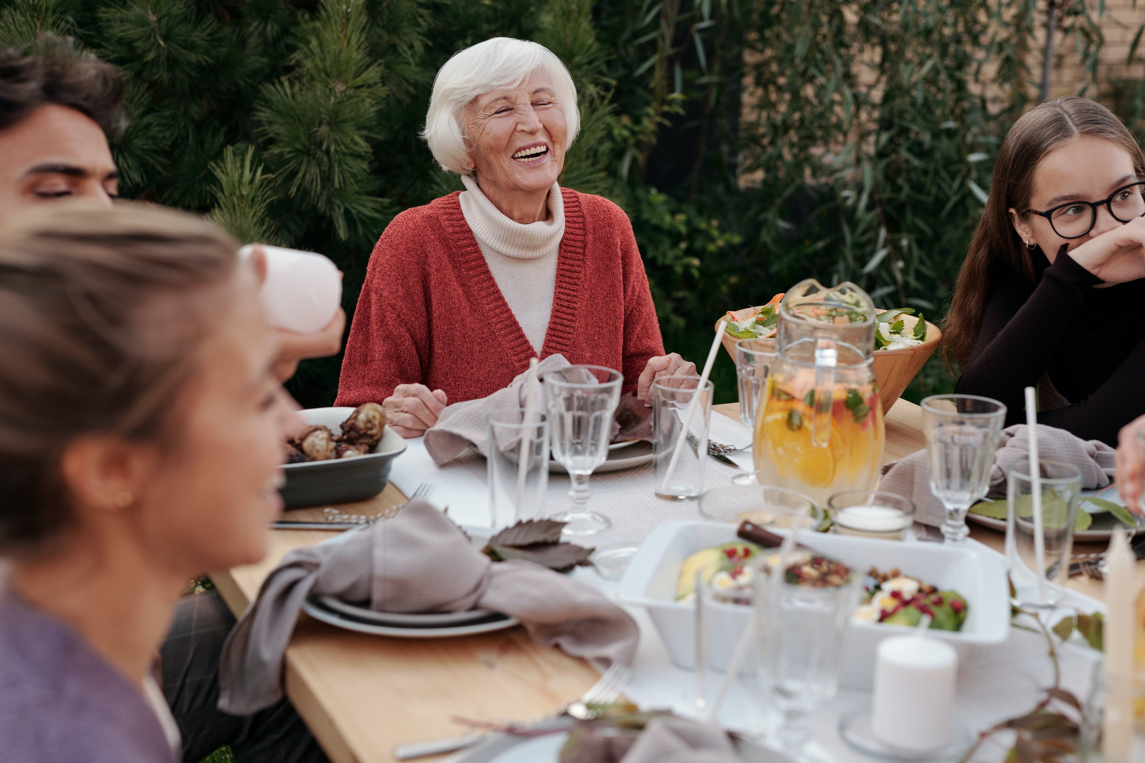 Group of people enjoying a meal outdoors, with an elderly woman in a red sweater laughing at the center of the table. - Home Instead