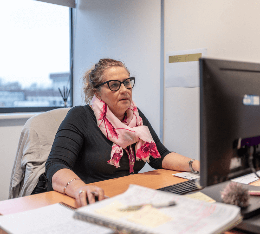 Person wearing glasses and a pink scarf, working on a computer at a desk in an office setting. - Home Instead