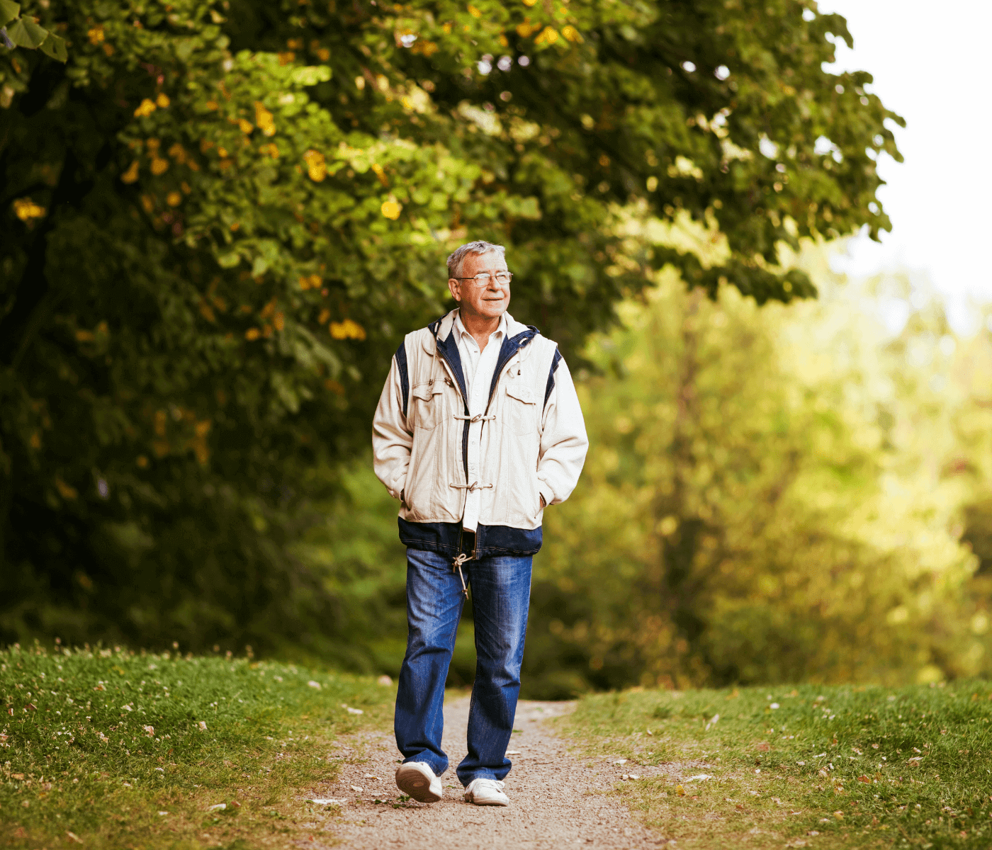 A man in a beige jacket and jeans walks along a path in a lush, green park, hands in pockets, looking content. - Home Instead