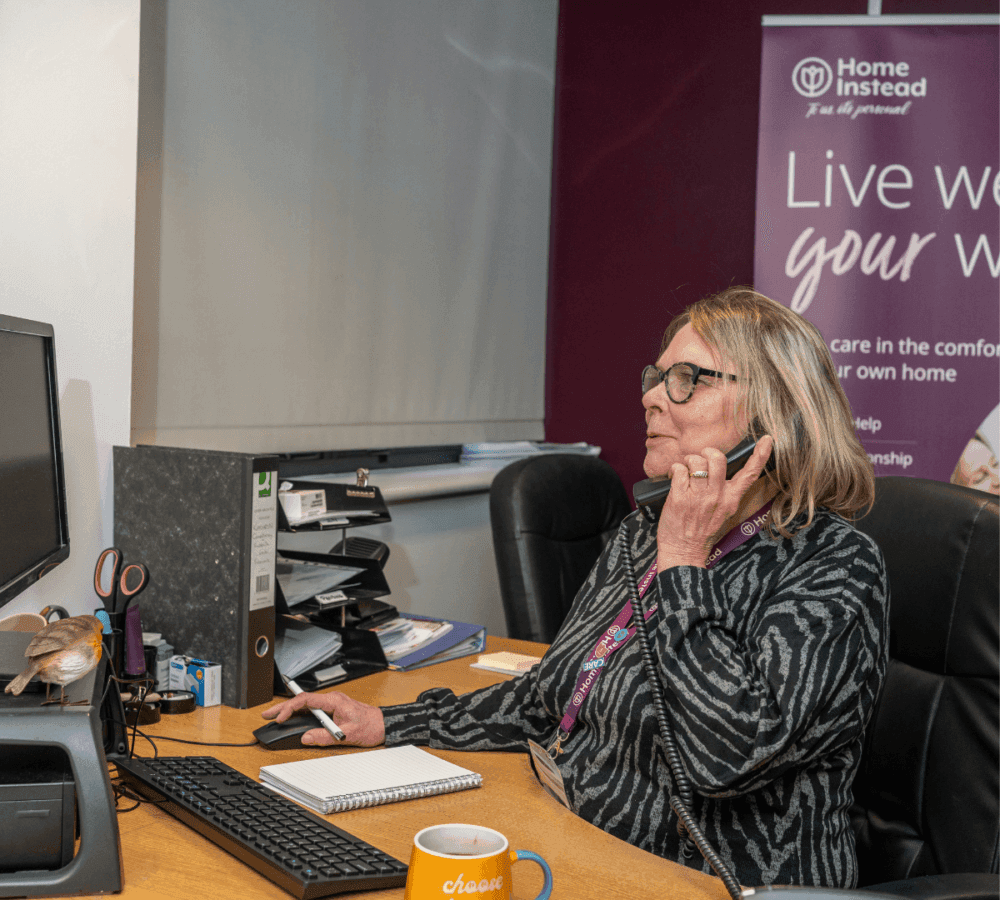 Woman at a desk on the phone, with a computer mouse in hand and a bird on the desk. Poster with text behind her. - Home Instead