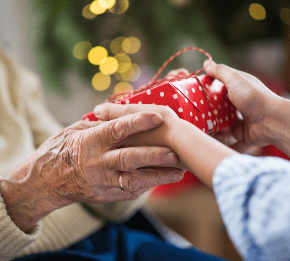 Hands of a young person and an elderly person holding a wrapped, red polka-dot present, with festive lights in background. - Home Instead