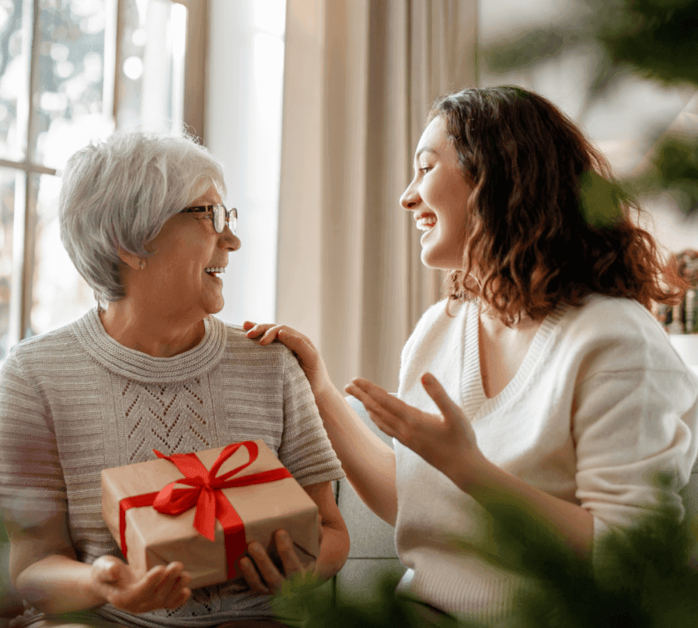 Elderly woman receiving a wrapped gift with a red ribbon from a smiling young woman inside a cozy room. - Home Instead
