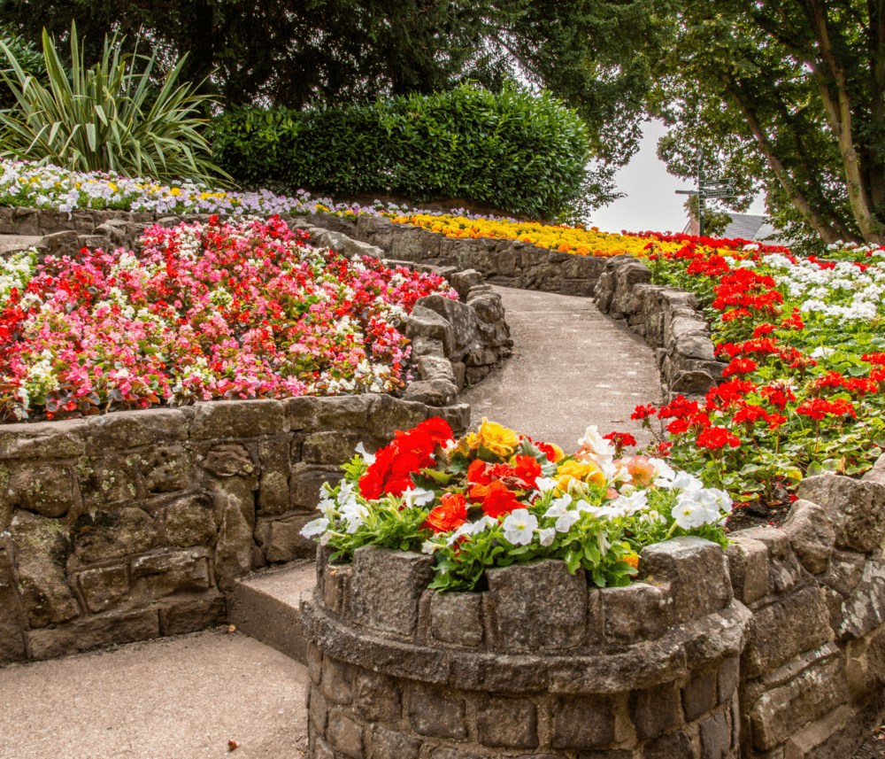 Stone path through a colorful flower garden with red, yellow, pink, and white blooms, surrounded by lush greenery. - Home Instead