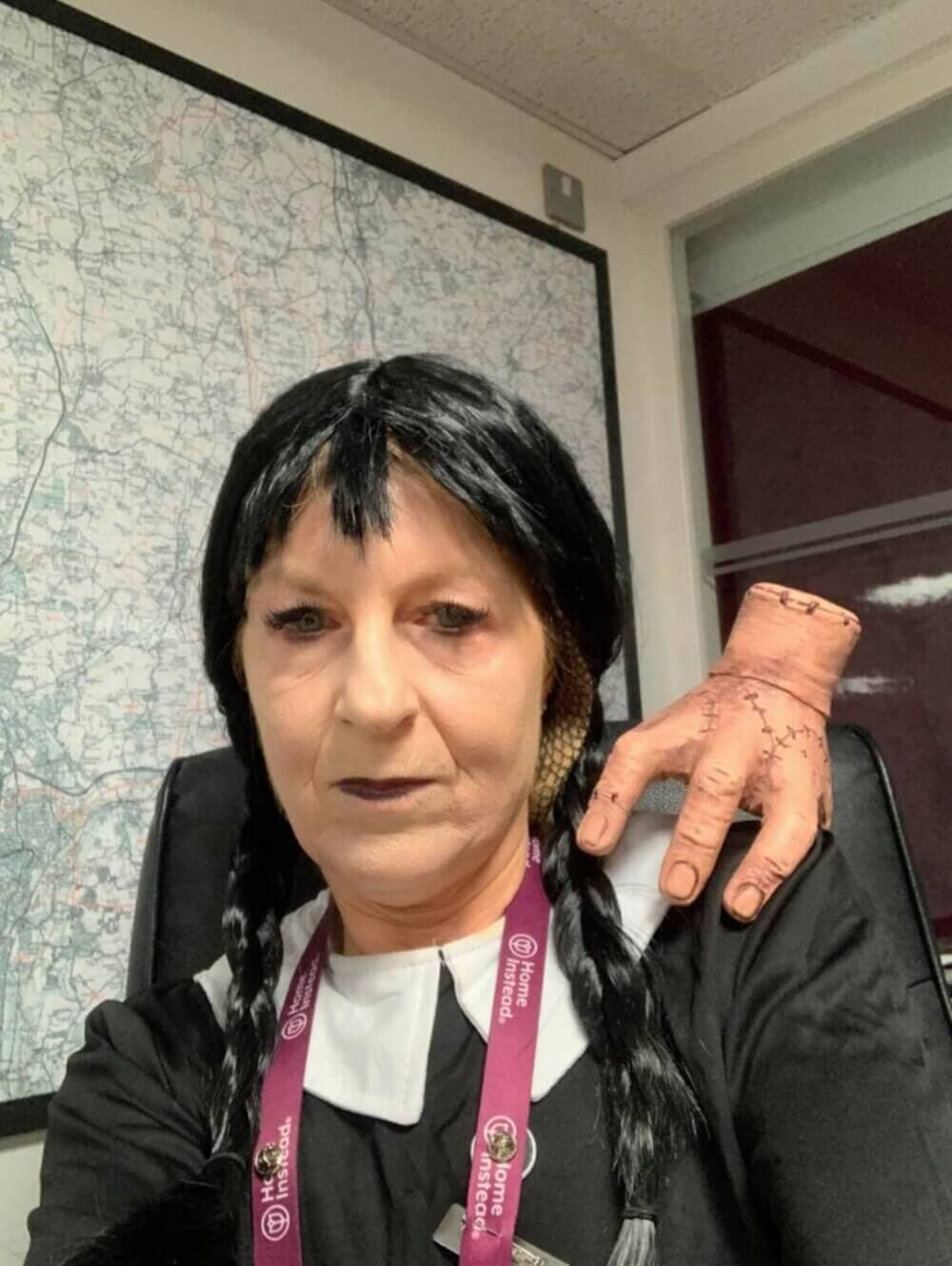 Person dressed as a fictional character with black braided hair, pale face, and a fake hand on their shoulder. - Home Instead