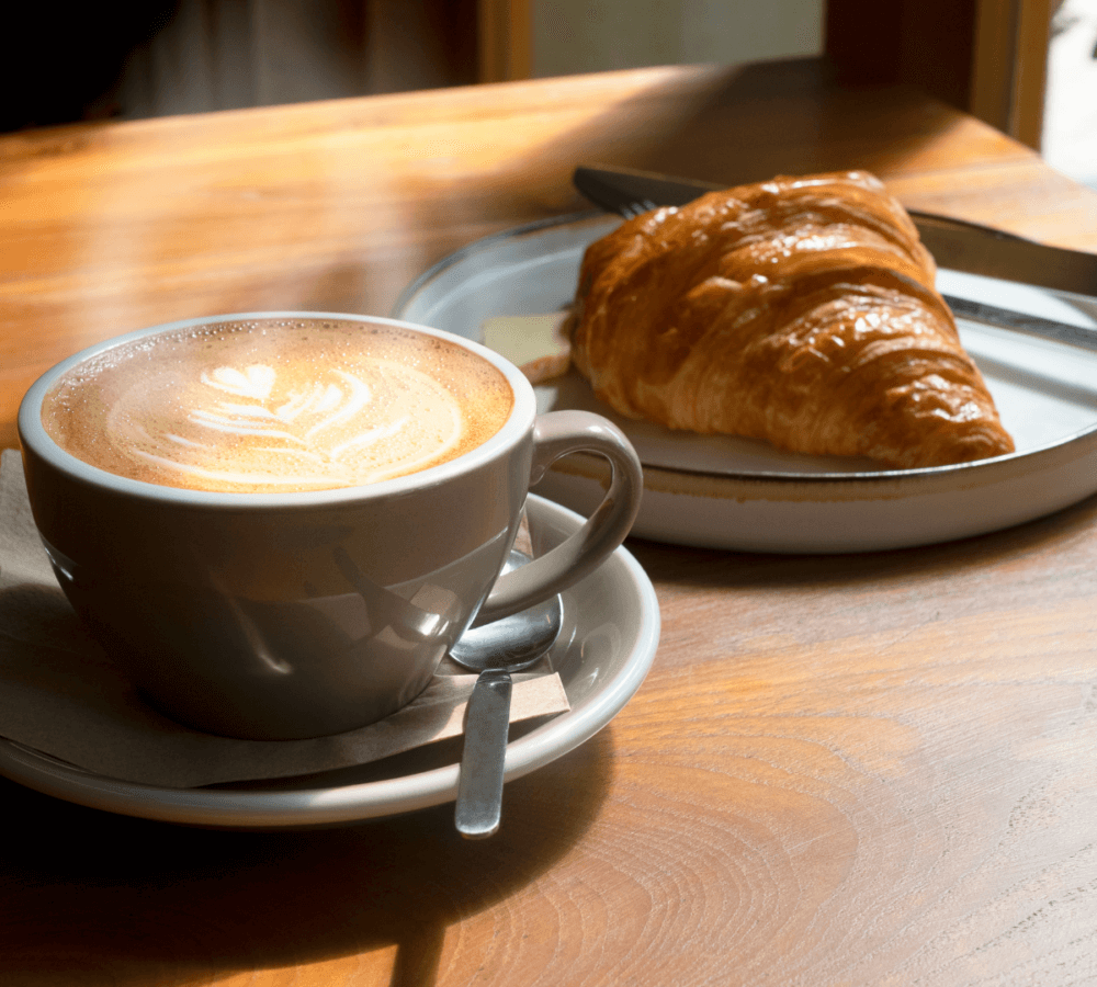 A cup of latte with latte art on saucer and spoon next to a plate with a croissant on a wooden table in sunlight. - Home Instead