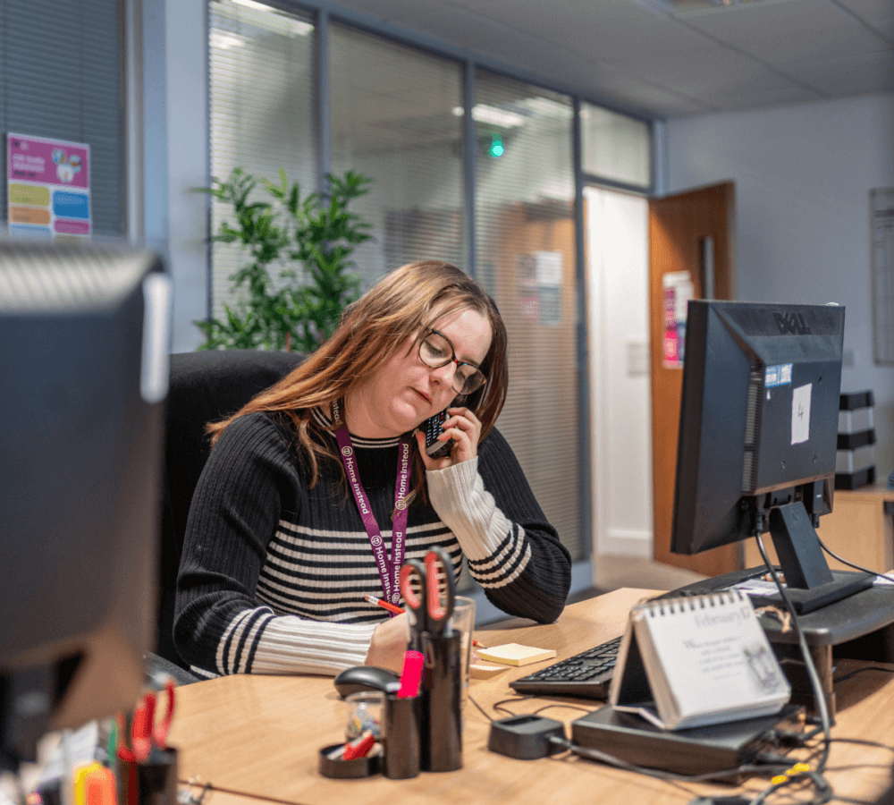 Woman at office desk on phone, writing in notepad, with computer and office supplies on desk, background with plants. - Home Instead