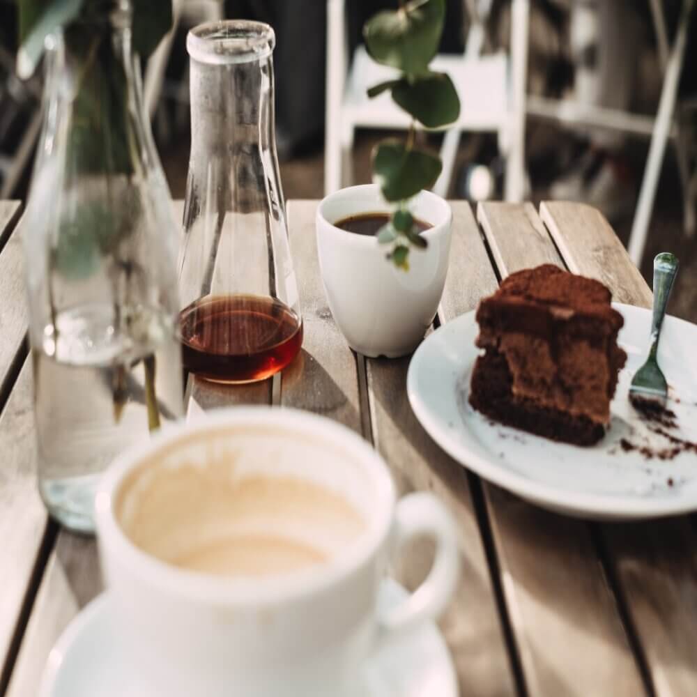 A wooden table with coffee, a glass bottle, a white cup, a carafe of coffee, and a plate of chocolate cake. - Home Instead
