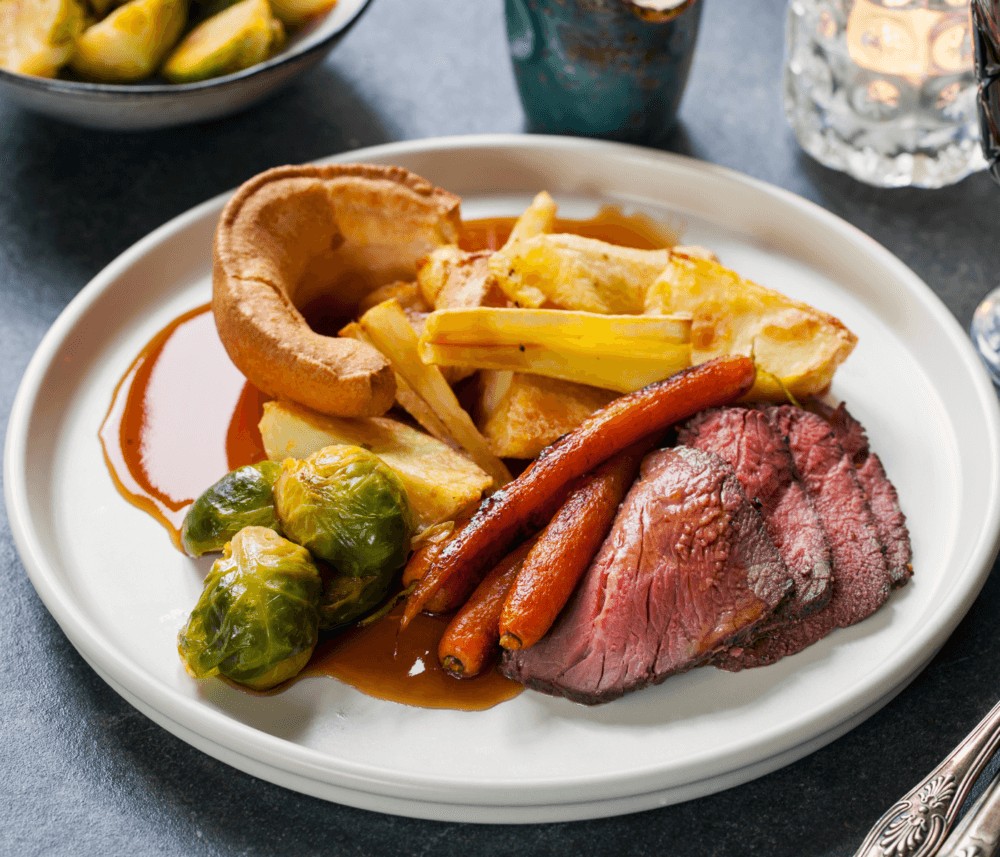 A plate with roast beef, carrots, potatoes, Brussels sprouts, and Yorkshire pudding, served with gravy. - Home Instead