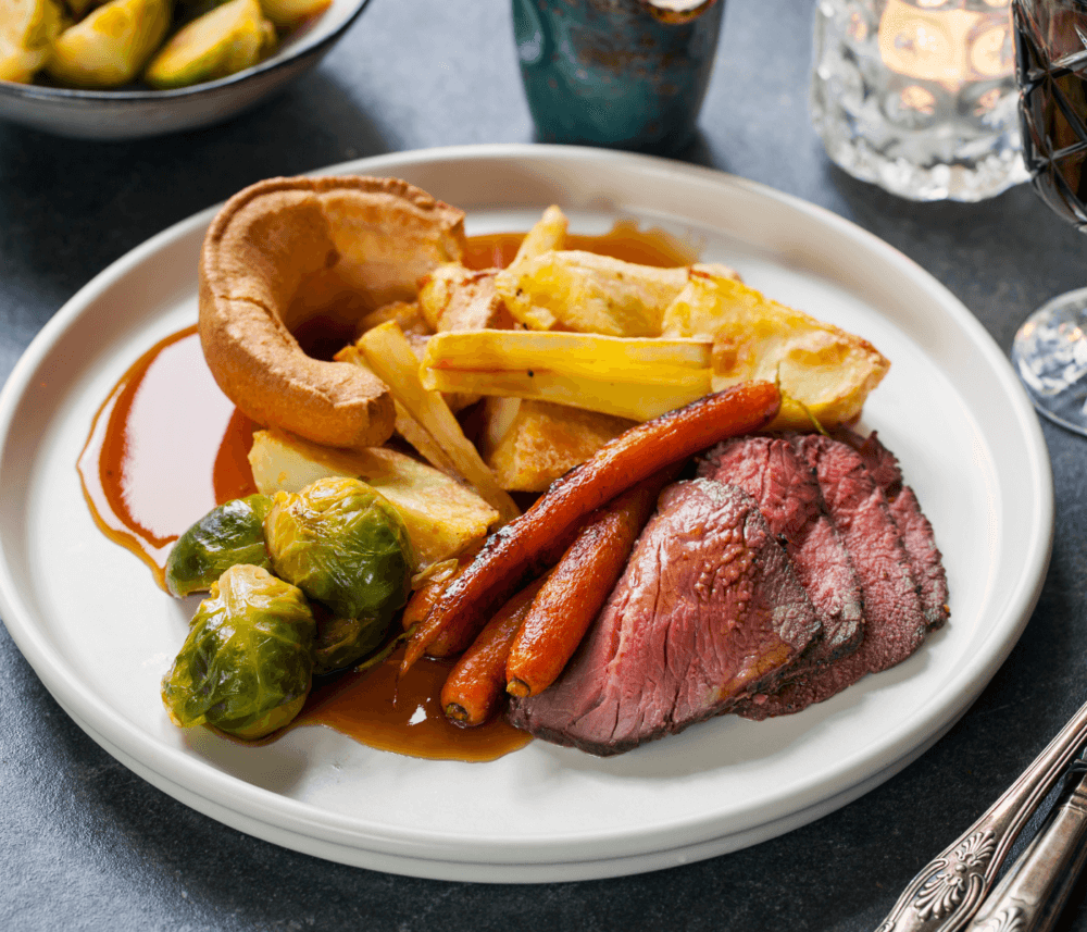 A plate of roast beef, carrots, brussels sprouts, parsnips, Yorkshire pudding, and gravy on a dining table. - Home Instead