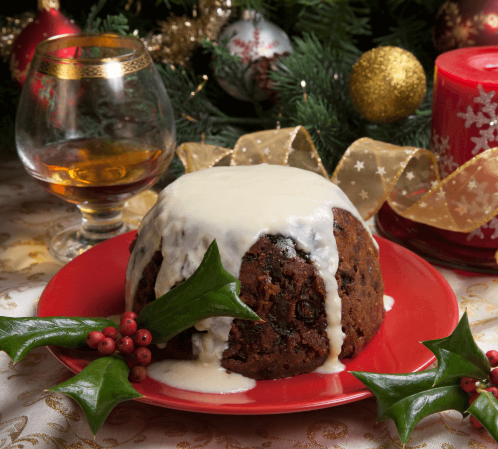 Christmas pudding with white sauce on a red plate, holly garnish, brandy glass, and festive decorations in the background. - Home Instead