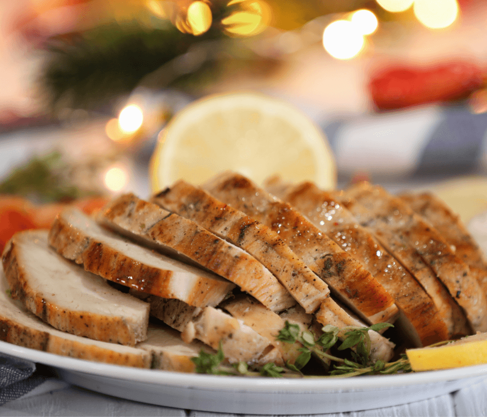 Sliced roasted turkey breast on a white plate, garnished with herbs, with festive lights blurred in the background. - Home Instead