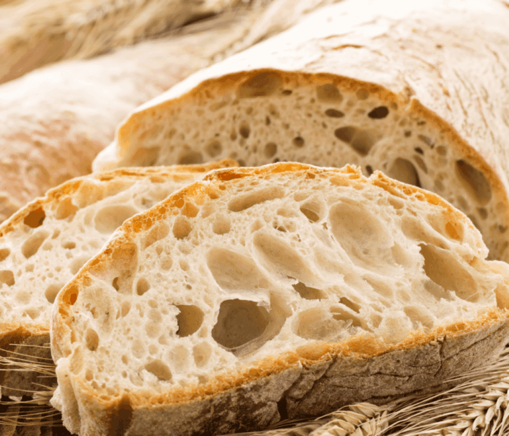 Slices of fresh, rustic bread with a crusty exterior and airy interior placed on a bed of wheat stalks. - Home Instead