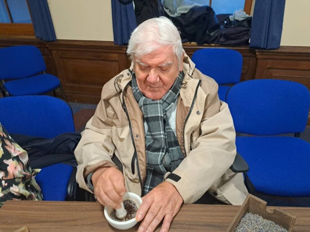 An elderly man in a beige coat and scarf sits at a table, looking down at a small bowl filled with herbs. - Home Instead