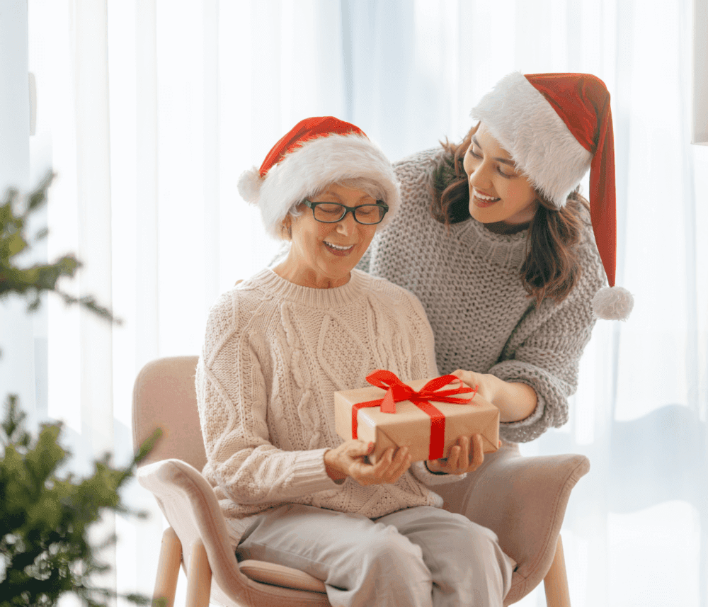 Two women in Santa hats smiling, the younger one giving a wrapped gift to the older one, near a Christmas tree. - Home Instead