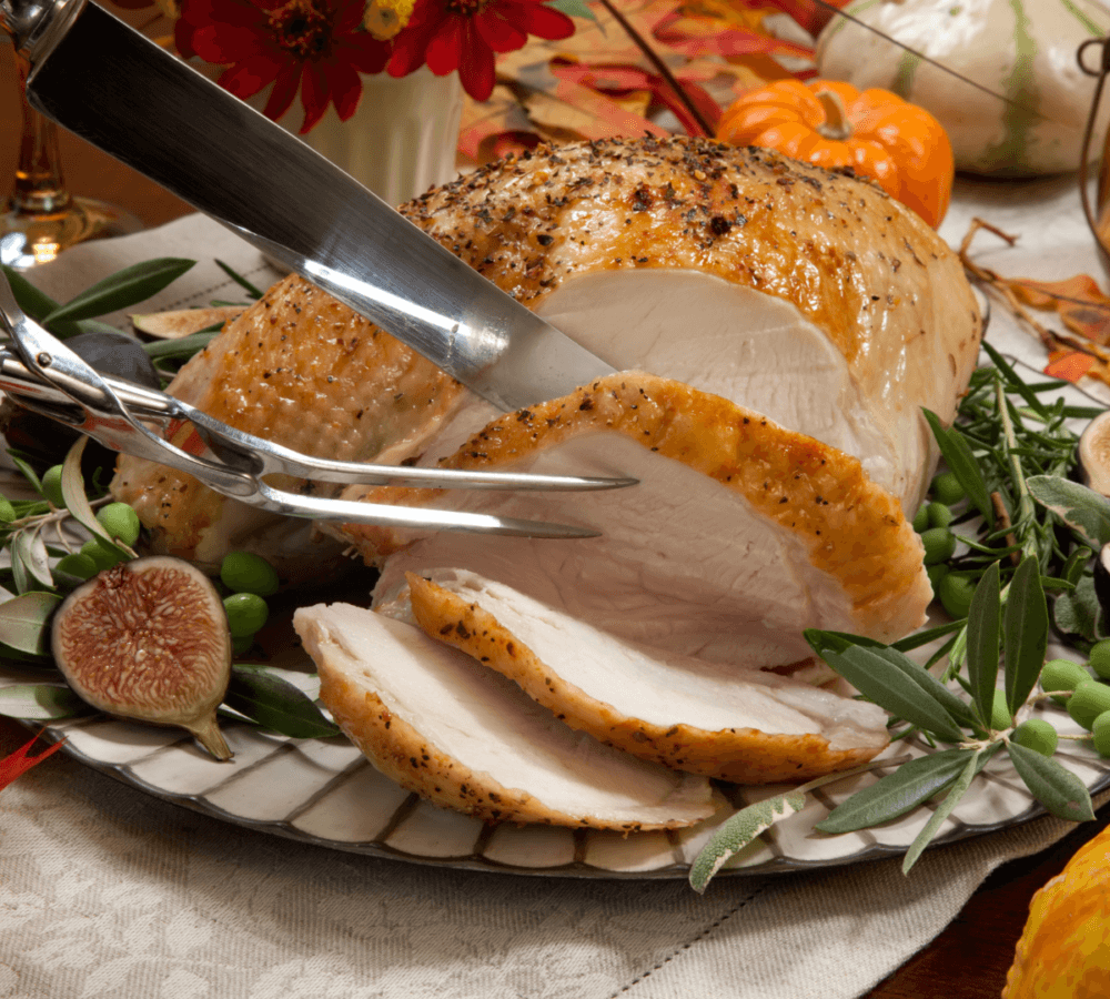 A knife and fork carving slices from a roasted turkey breast, garnished with figs and leafy greens, on a festive table. - Home Instead
