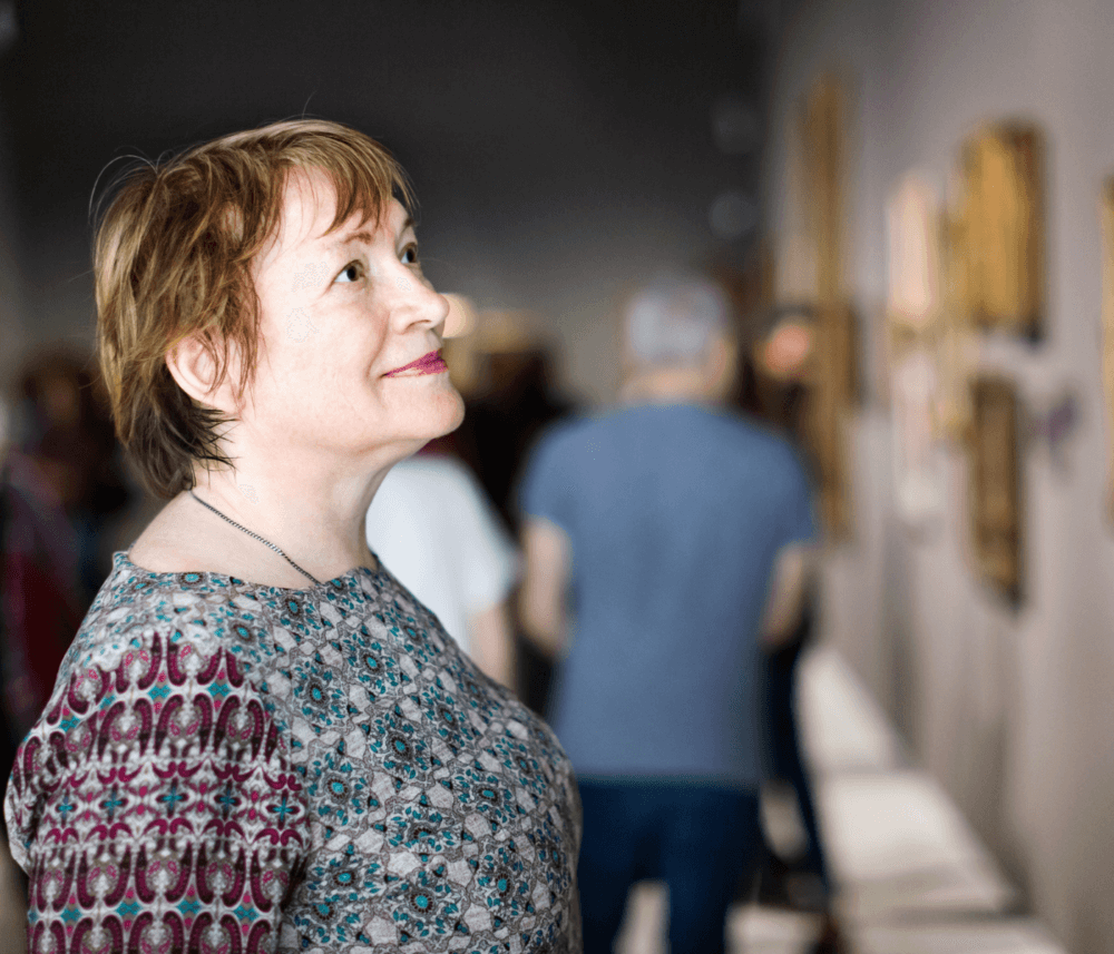 A woman with short hair and a patterned shirt looks at artwork in a gallery, surrounded by other visitors. - Home Instead