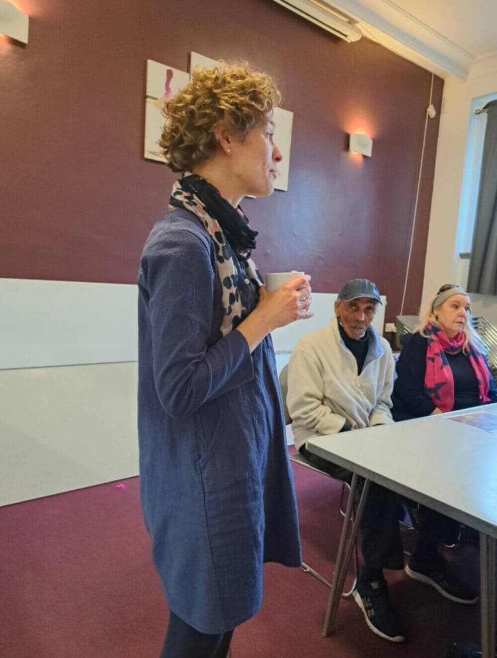 A person stands and speaks to two seated individuals in a room with maroon walls and artwork on the wall. - Home Instead