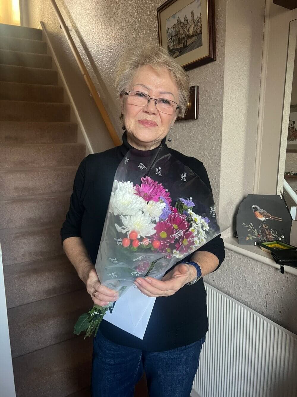 Elderly woman with short grey hair holding a colorful bouquet of flowers stands inside a house near stairs. - Home Instead