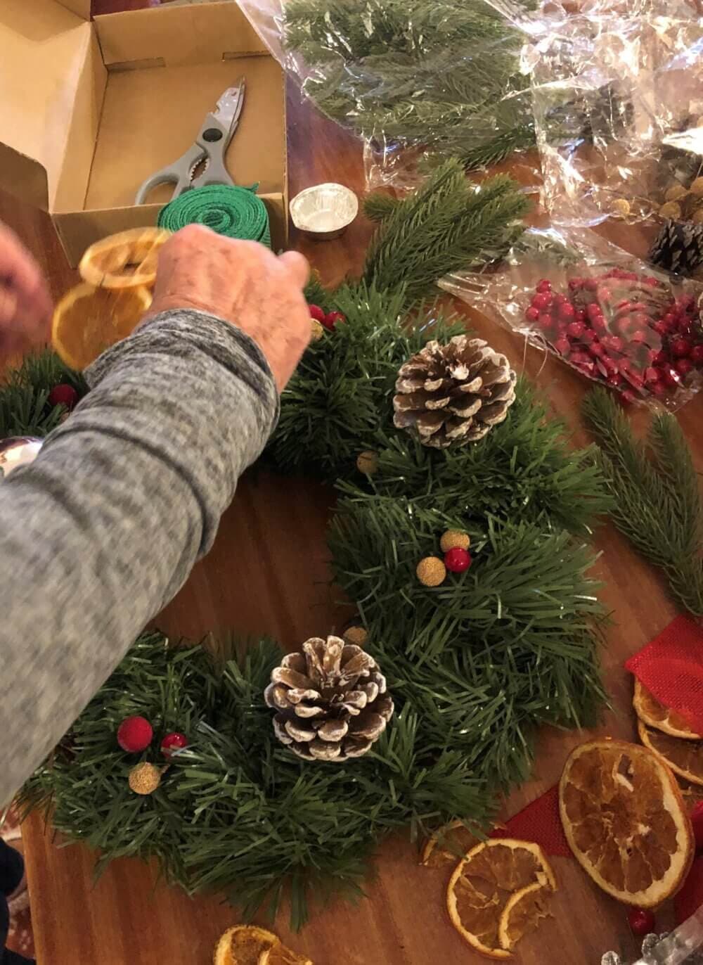 Hands decorating a Christmas wreath with dried orange slices, pine cones, and berries on a wooden table. - Home Instead