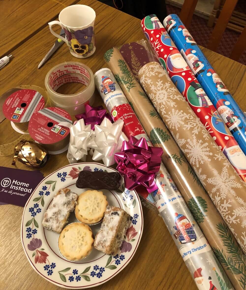Wrapping paper rolls, ribbons, tape, a mug, a plate with snacks, and a booklet on a wooden table. - Home Instead