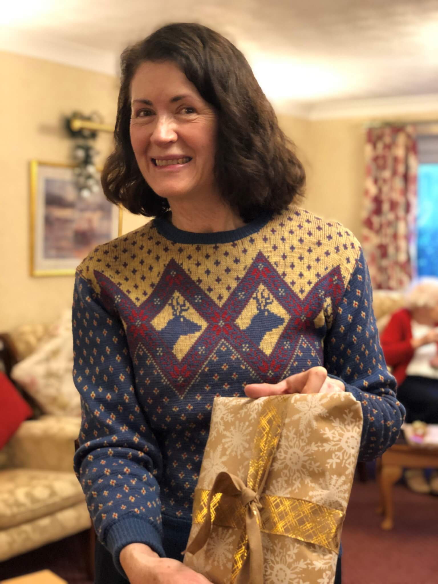 Woman smiling and holding a wrapped gift in a decorated living room during the holiday season. - Home Instead