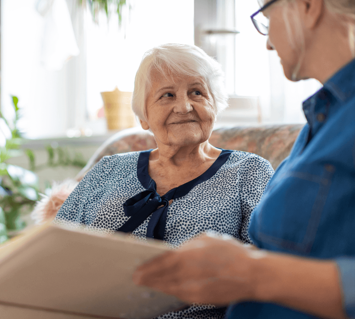 An elderly woman and a younger woman sit together, with the elderly woman looking at the younger one holding a book. - Home Instead
