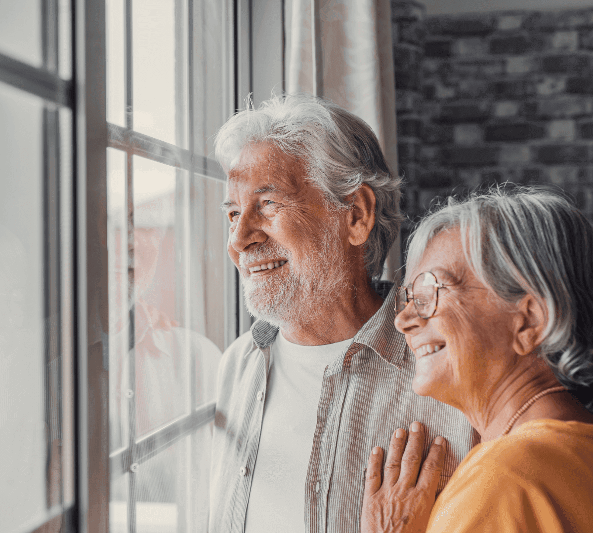 An elderly couple smiling and looking out of a window; woman with glasses and man with white beard. - Home Instead