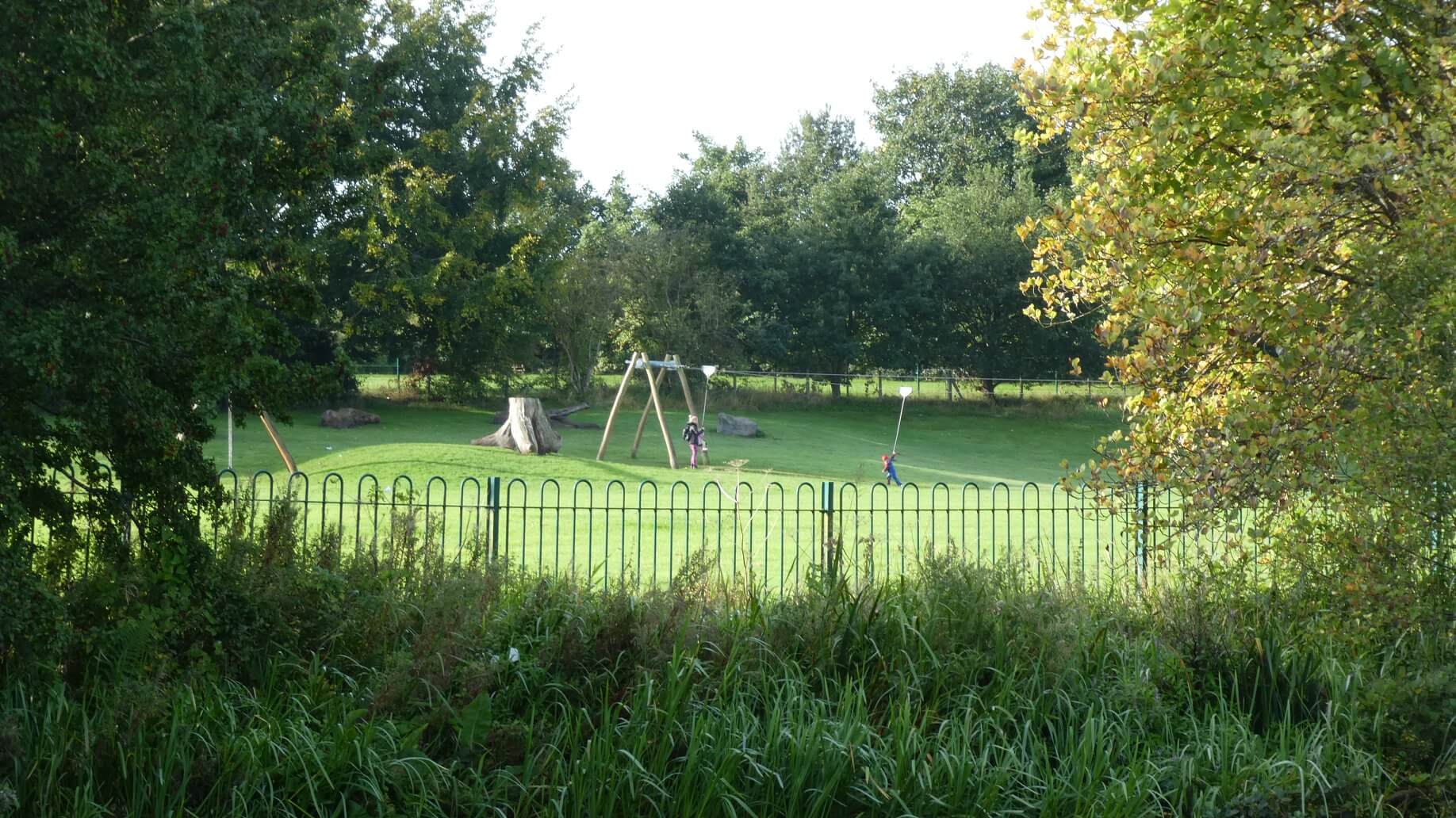 A playground with swings, surrounded by trees and green grass, seen through a wrought-iron fence on a sunny day. - Home Instead