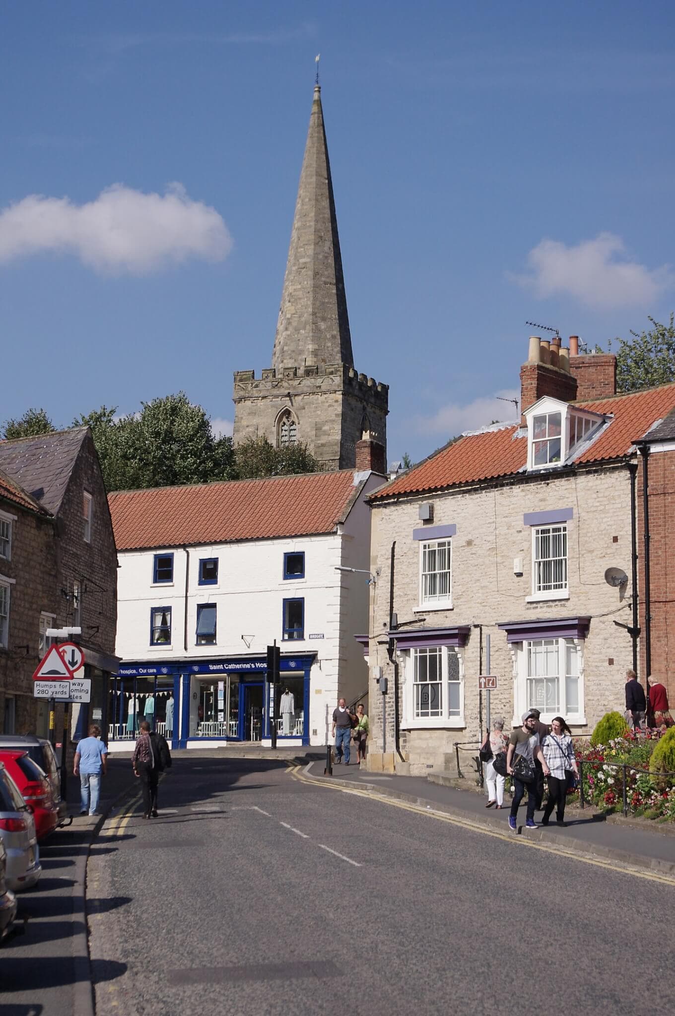 A street scene with a stone church spire in the background, people walking, and cars parked along the road. - Home Instead