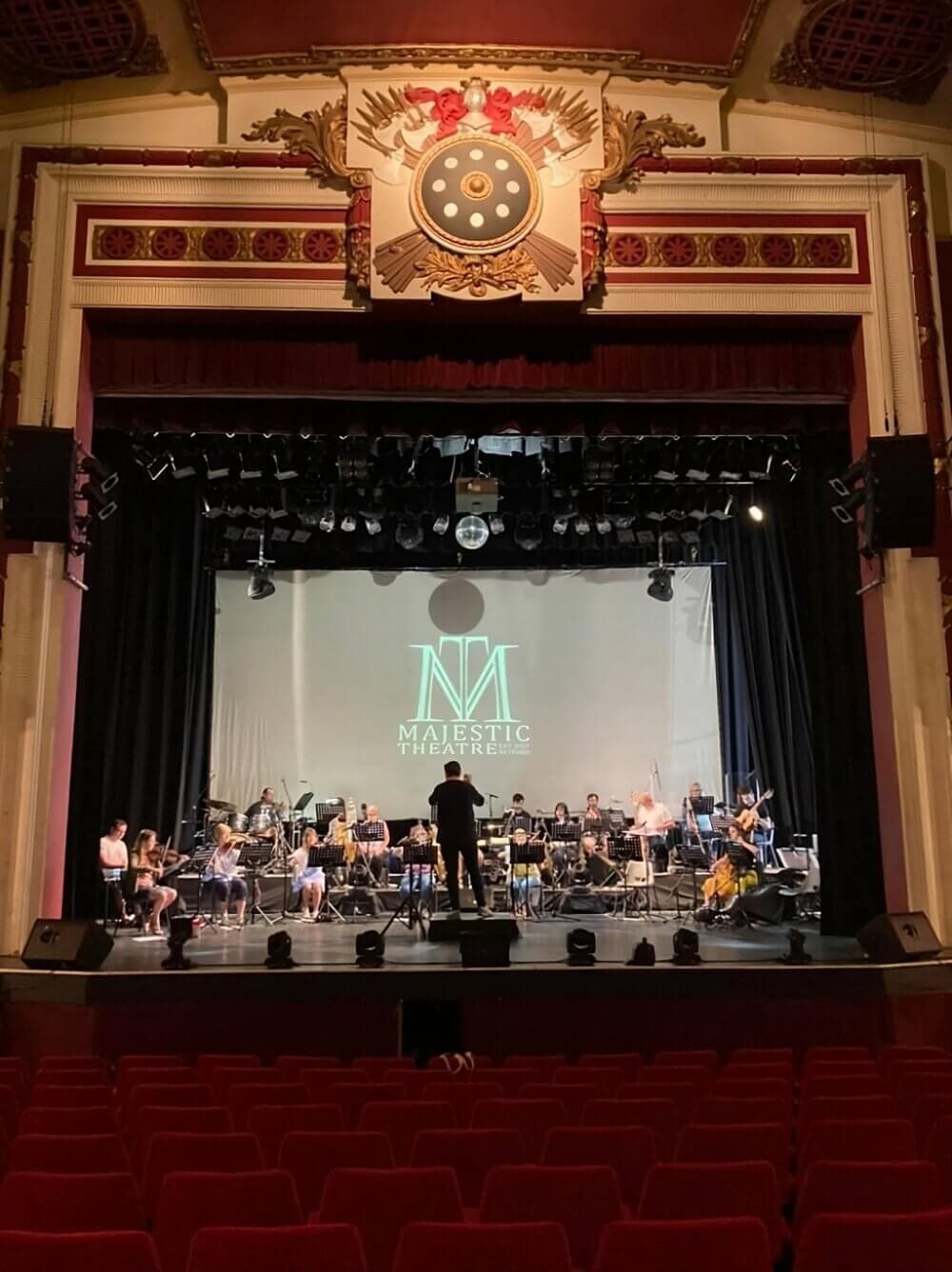 An orchestra rehearses on a stage with a conductor, in a theater with red seats and a "Majestic Theater" logo on screen. - Home Instead