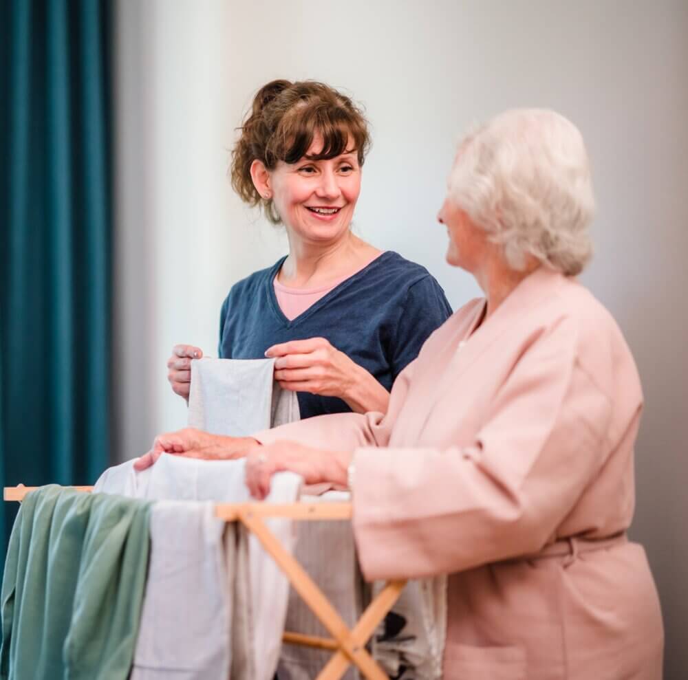 A young woman and an older woman smile while doing laundry together. The younger woman holds a piece of clothing. - Home Instead