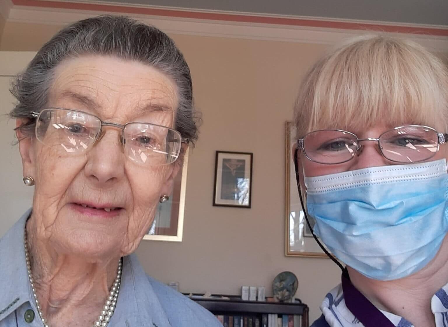 An elderly woman wearing a pearl necklace and a younger woman with glasses and a face mask taking a selfie. - Home Instead