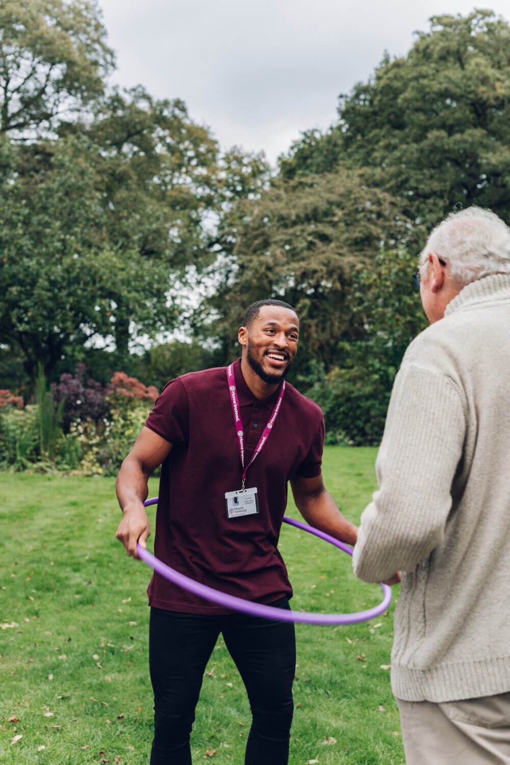 A man with a purple hula hoop smiles while interacting with an older man outdoors. - Home Instead