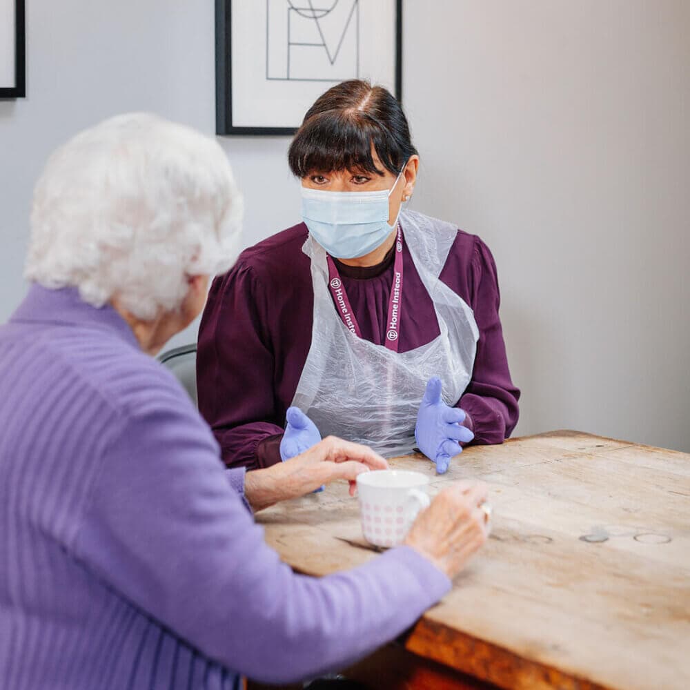A caregiver wearing a mask, gloves, and a plastic apron talks to an elderly woman at a wooden table. - Home Instead