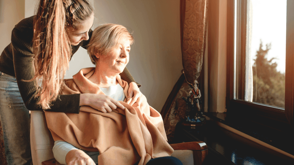 A young woman embraces an elderly woman sitting by a window with warm light streaming in. - Home Instead