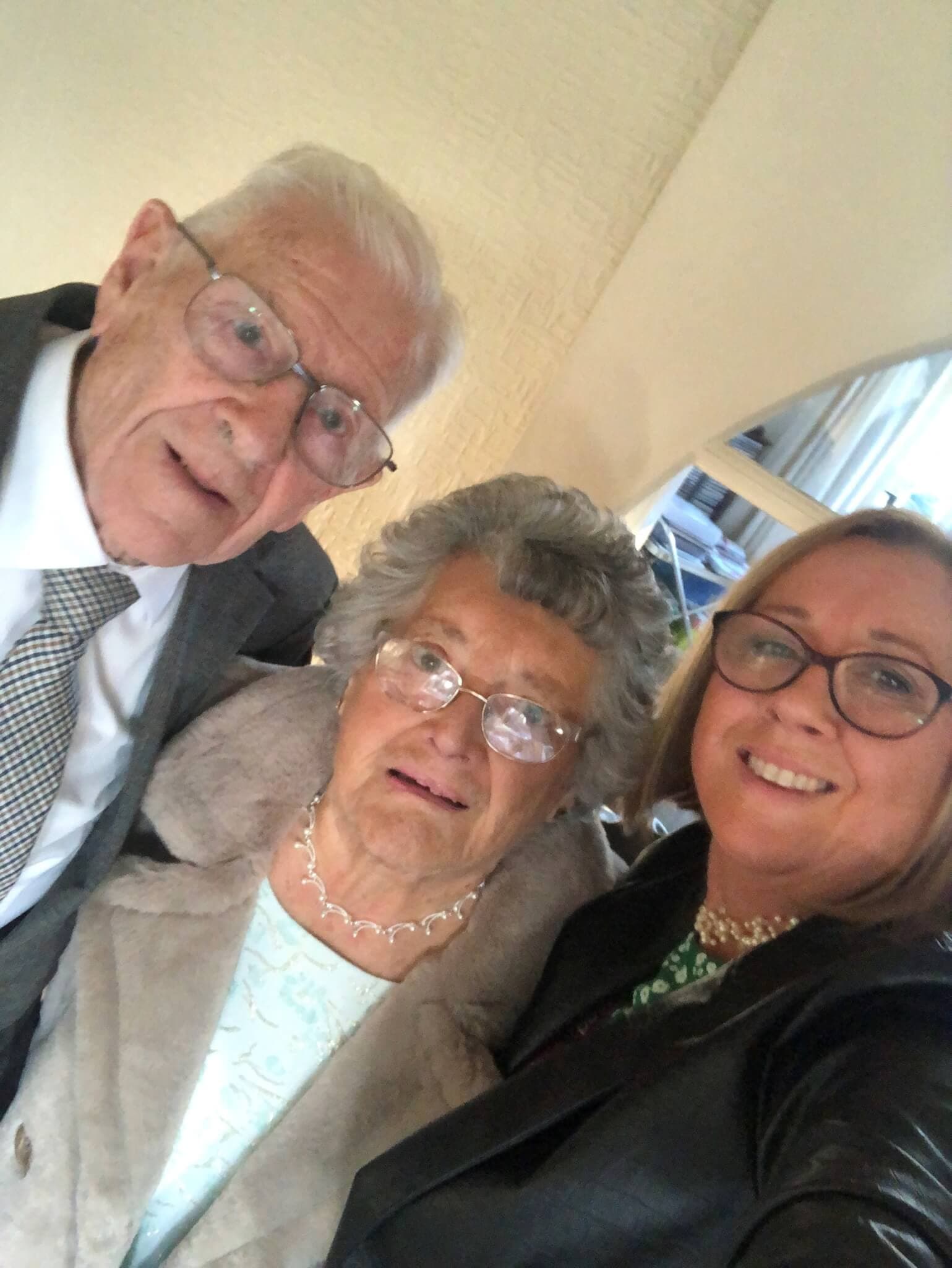 Three people, an elderly couple and a younger woman, smiling and looking at the camera indoors. - Home Instead