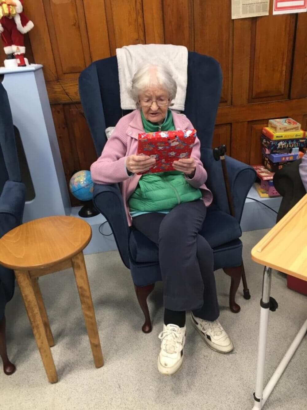 An elderly woman in a green and pink outfit sits in a chair, holding a wrapped gift. A side table and globe are nearby. - Home Instead